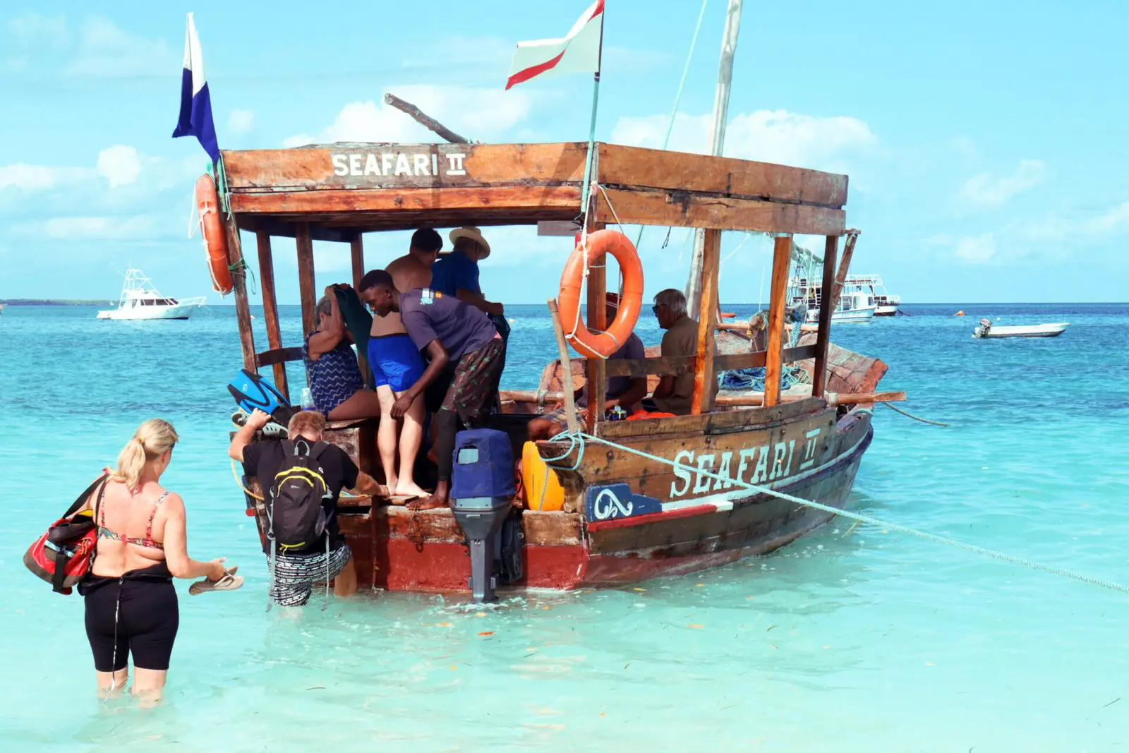 Guests wading through turquoise shallows to board a wooden dhow with a shaded roof deck in Zanzibar