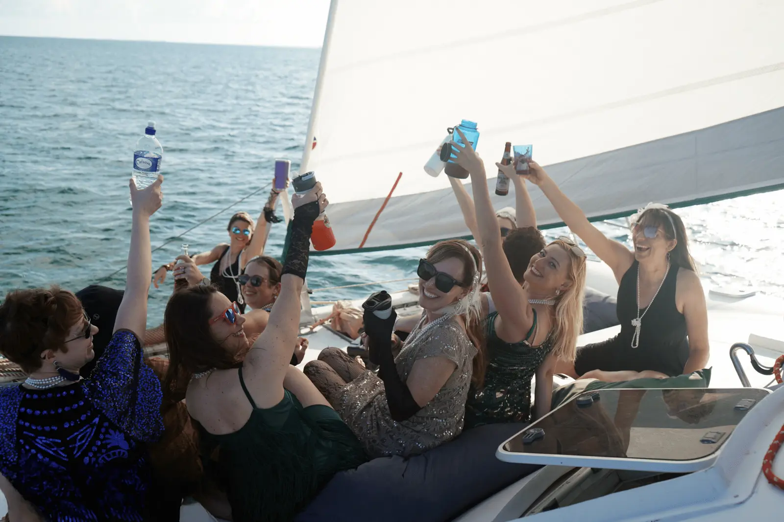 Women raising bottles and cups on a sailing catamaran deck beneath a full white sail