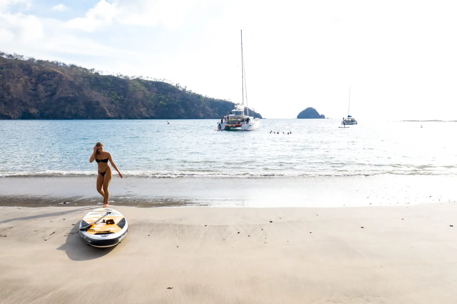 A woman walks up a quiet sandy beach in Papagayo past a beached paddleboard, sailboats offshore