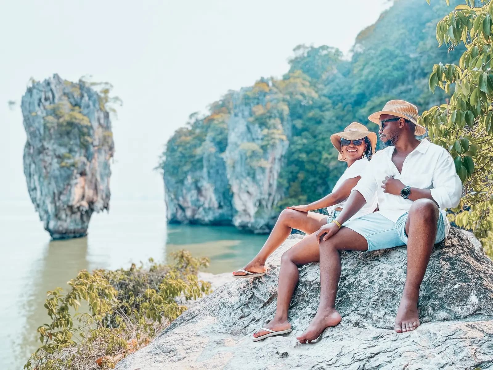 Couple in sun hats sitting on a rocky ledge facing the needle-shaped limestone rock in Phang Nga Bay