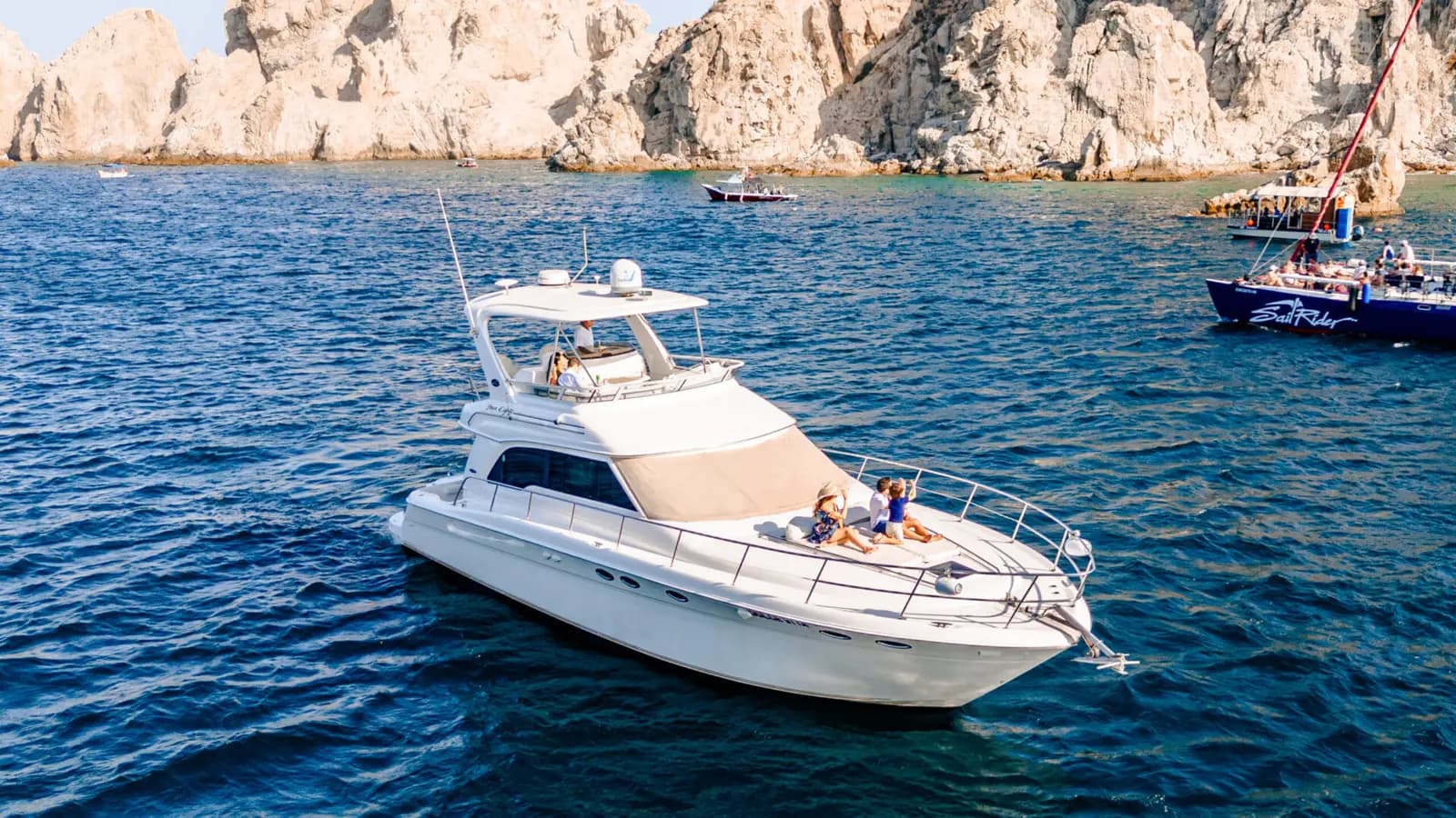 Flybridge yacht cruising deep blue water below rocky Los Cabos cliffs with other boats nearby