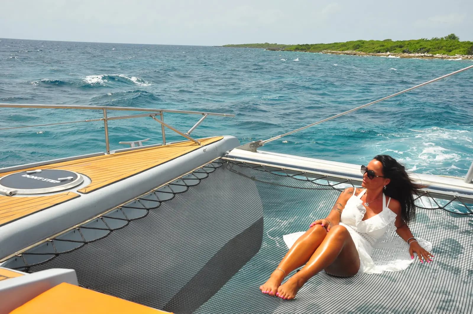 A woman in a white dress lounges on the catamaran's netting as it cruises past a green coastline