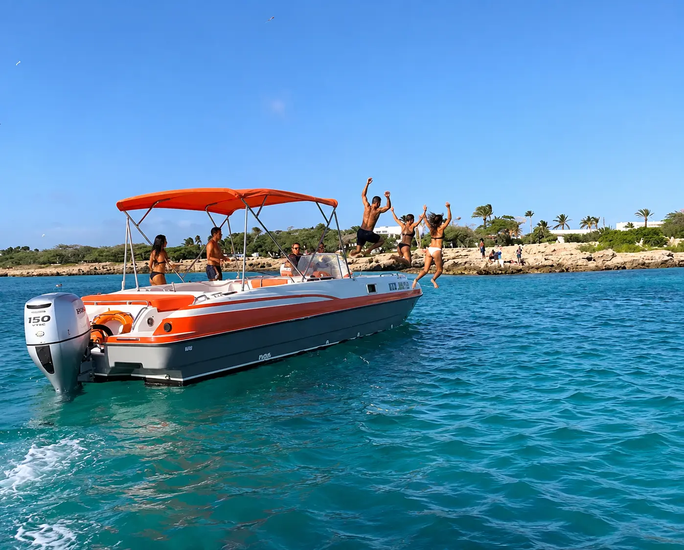 Guests jumping off the bow of a deck boat anchored near Aruba's rocky, palm-lined shore