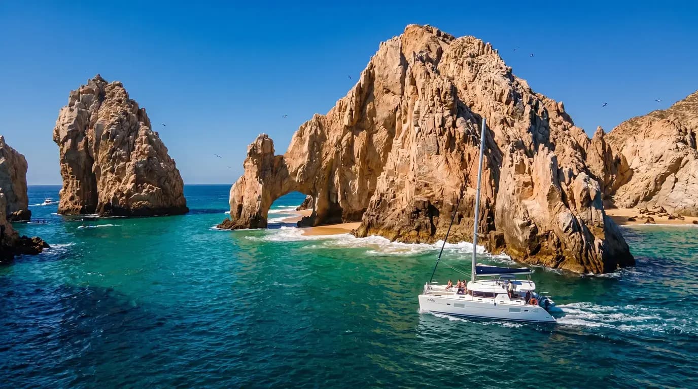 Catamaran with guests cruising past the rock arch and sea stacks of Cabo San Lucas in clear turquoise water
