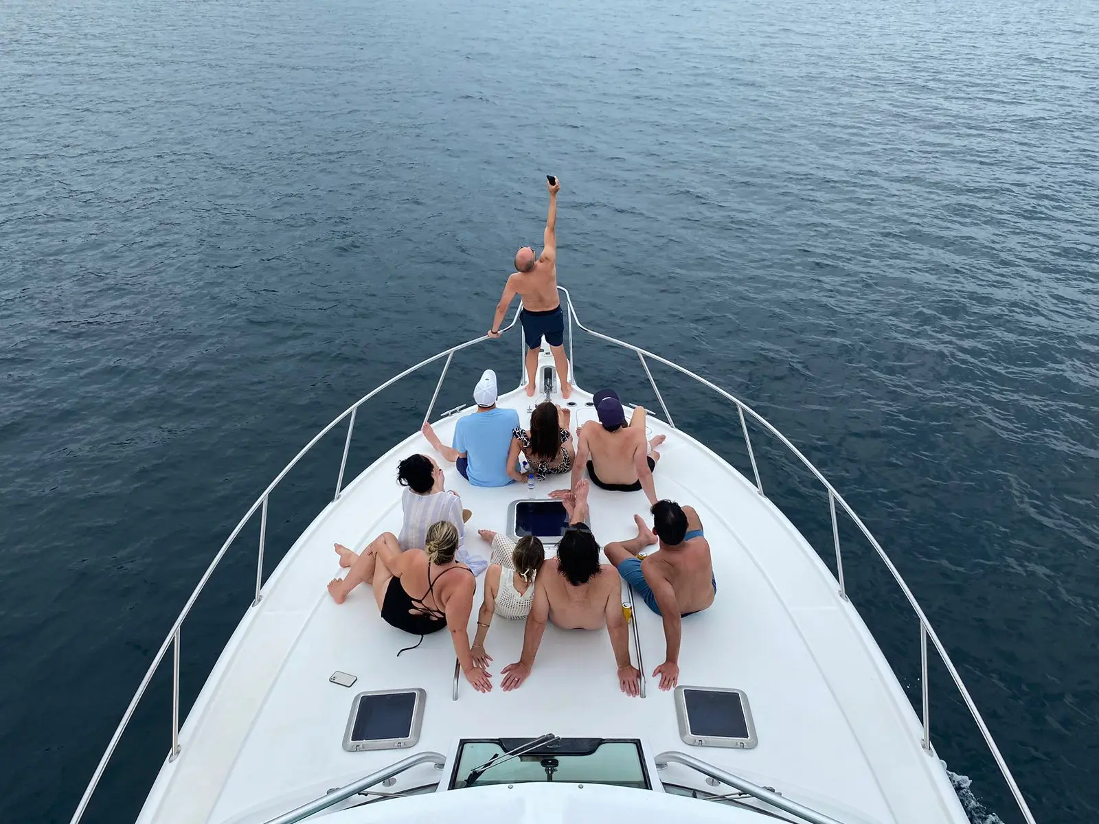 Seen from above, guests sit on the yacht's bow as a man stands at the tip with an arm raised