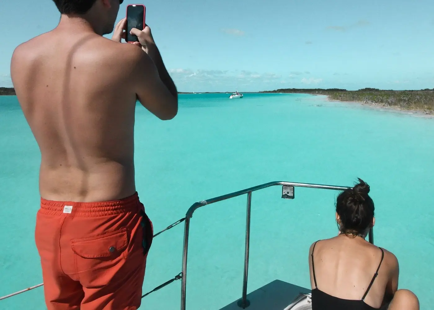 Man using his phone to capture Bacalar Lagoon from a catamaran bow as a woman relaxes