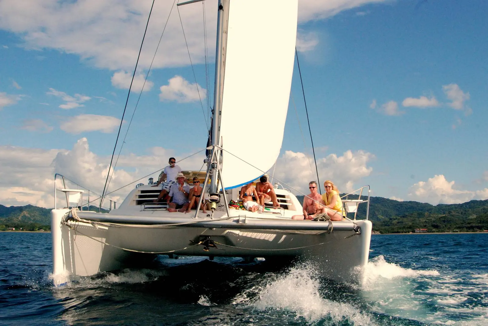 Guests riding on the bow of a sailing catamaran as it cuts through waves along Costa Rica's green coast