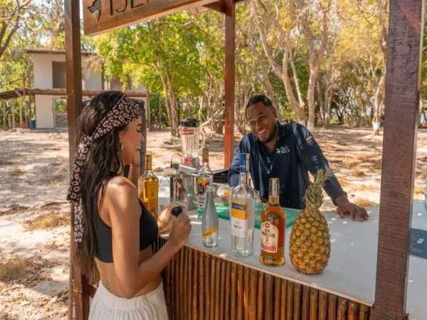 Woman chatting with a smiling bartender at a rustic wooden beach bar stocked with bottles and a pineapple