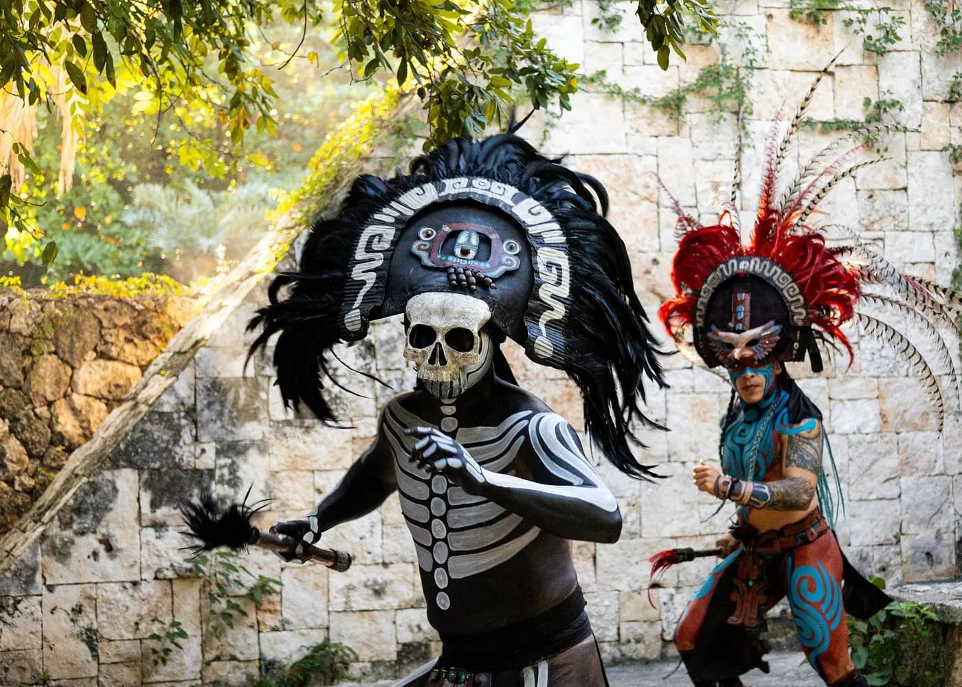 Two dancers in Mayan warrior costumes and feathered headdresses, one in skeleton body paint