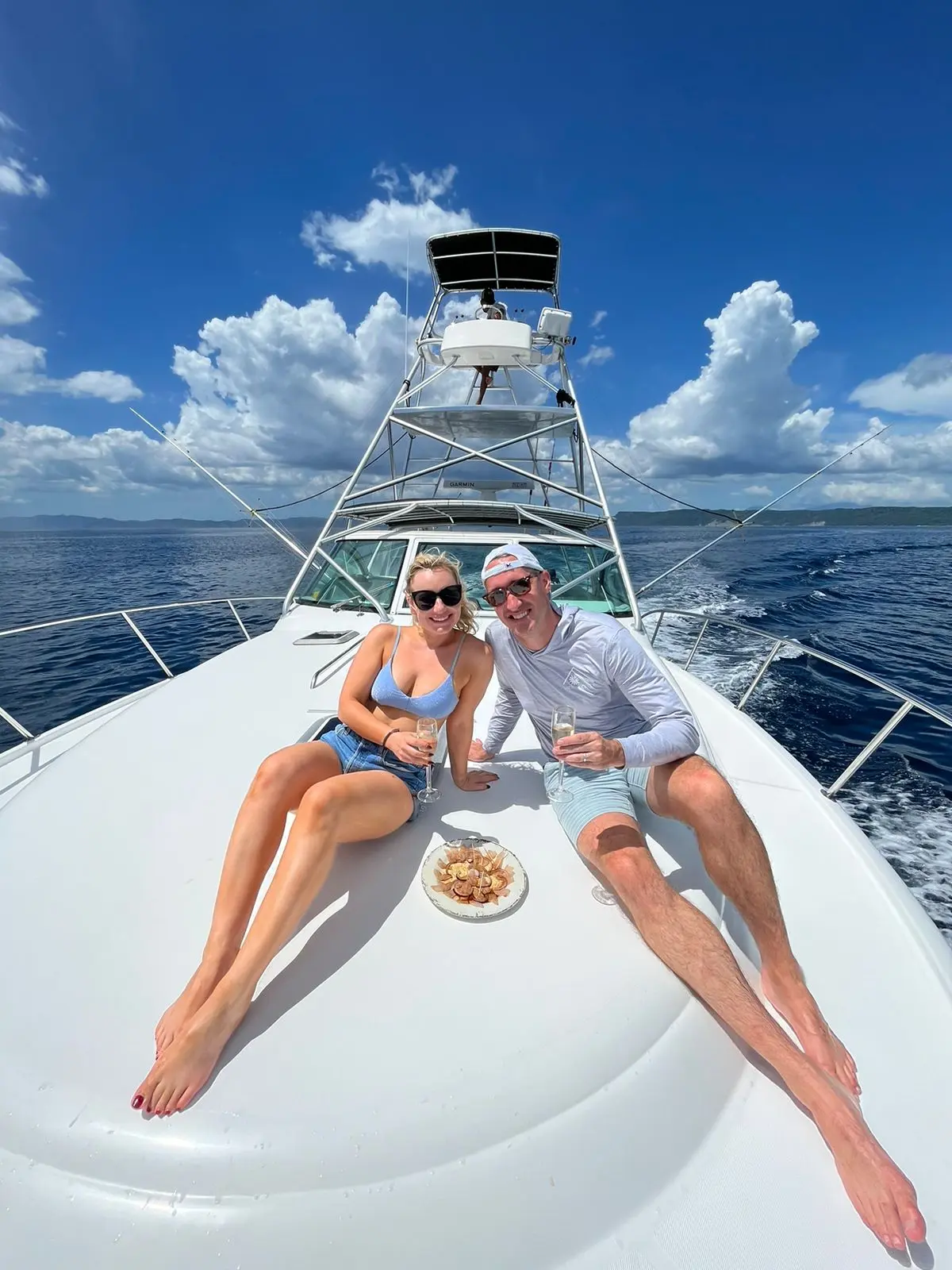 A couple relaxes on the bow of the sportfishing yacht with a plate of snacks under blue skies