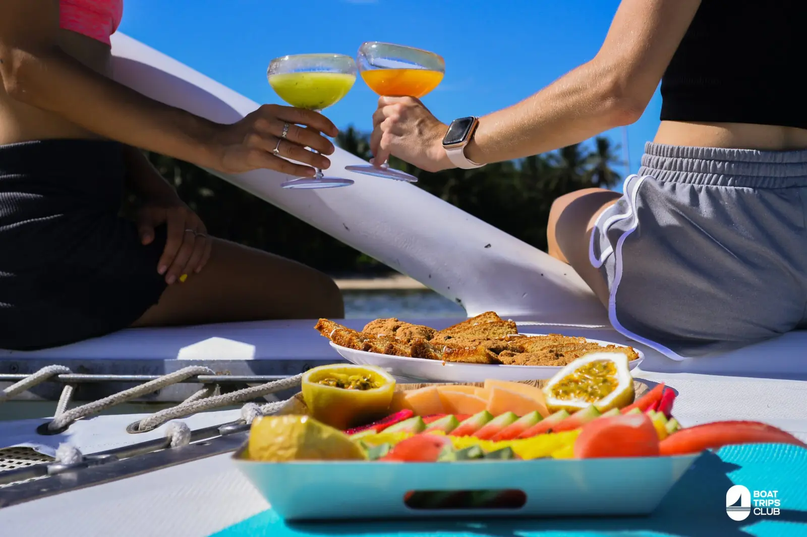 Two guests clink tropical cocktails over a platter of passion fruit, papaya and snacks on the catamaran