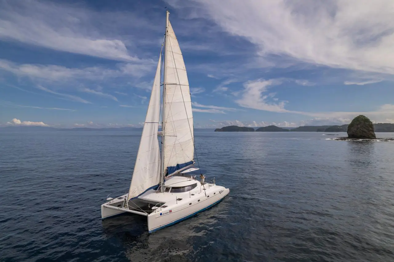 Sailing catamaran under full sail on calm water past a rocky islet off the Guanacaste coast