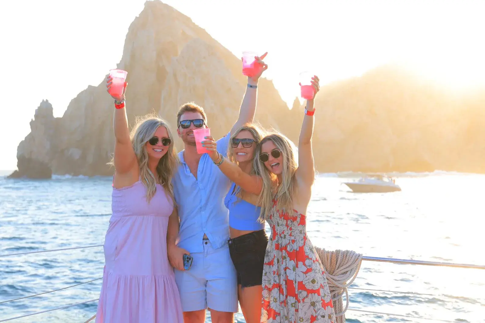 Four guests raise pink drinks on the catamaran as the sun sinks behind the Cabo San Lucas rocks