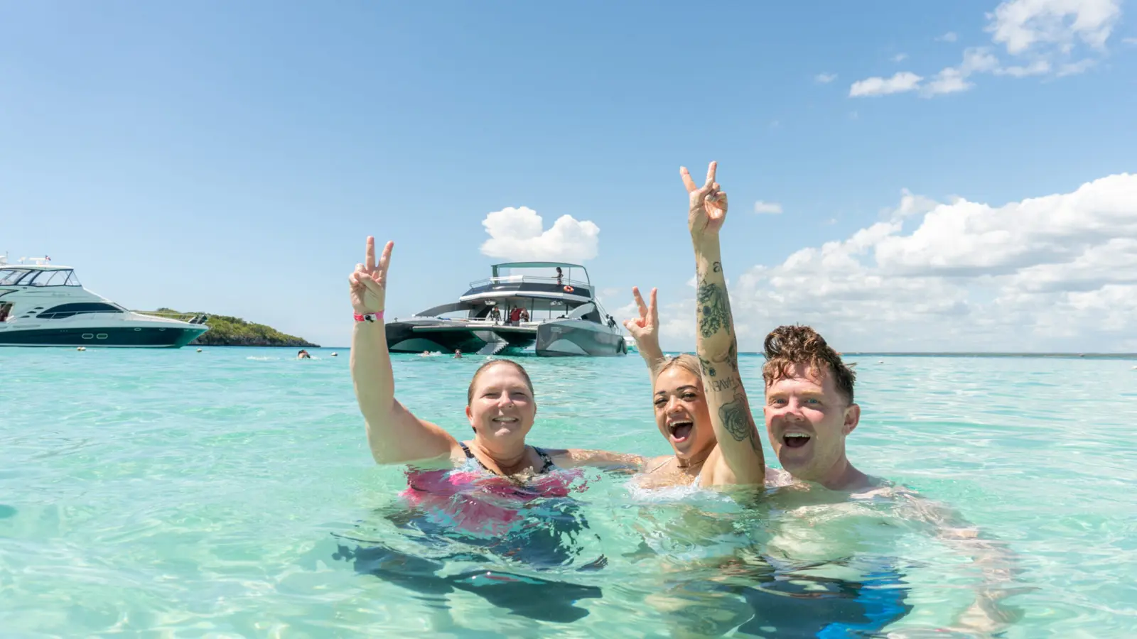 Three friends in turquoise water flashing peace signs, with two boats anchored behind them