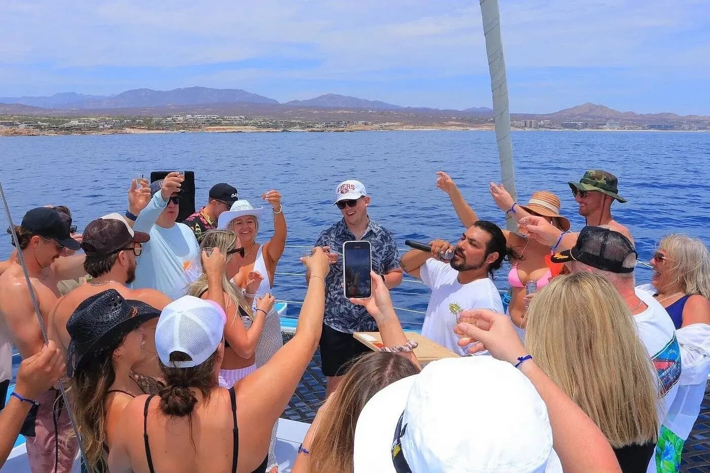 Guests raising cups and phones as they dance on a catamaran deck with mountains across the bay