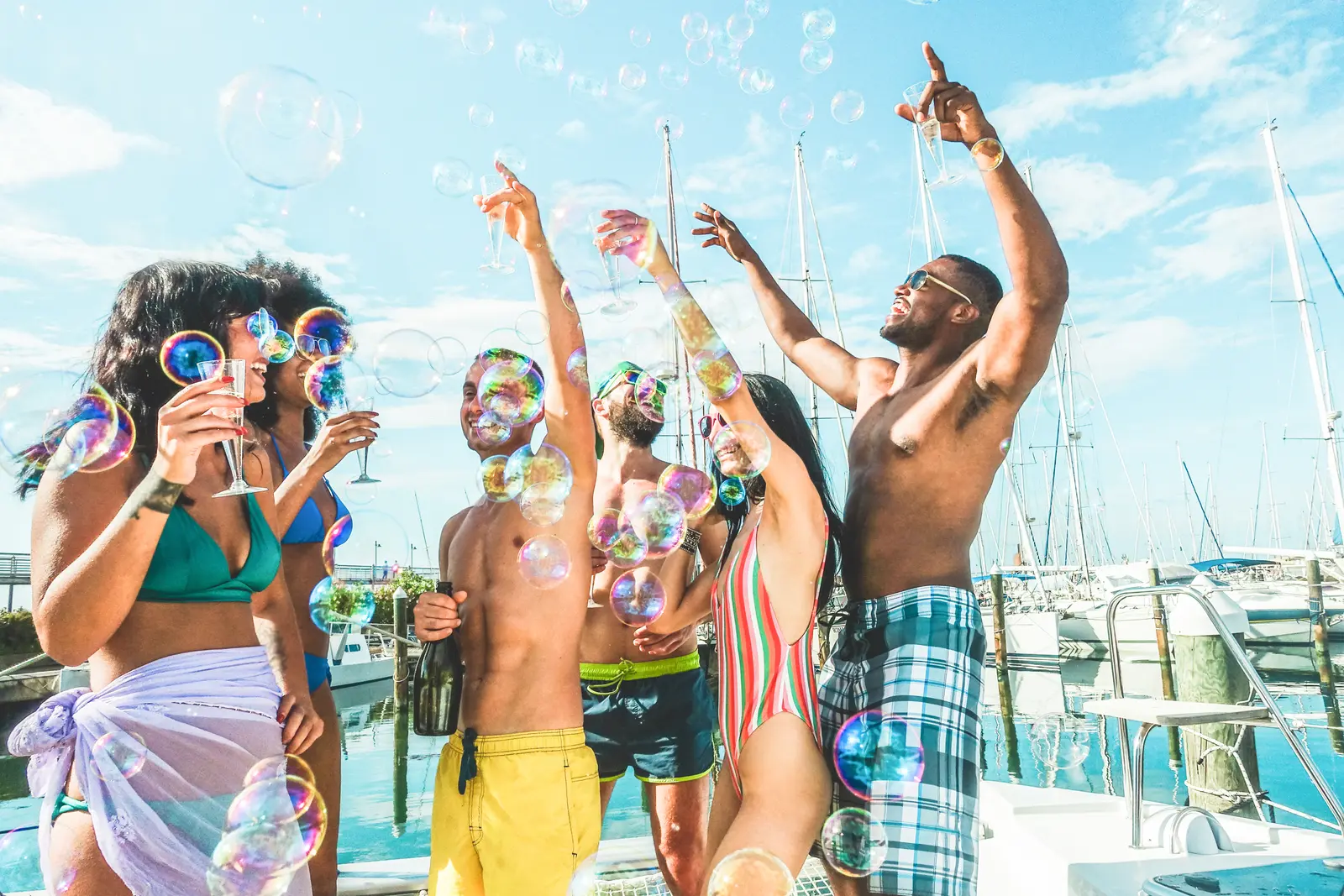 Group of friends raising their arms as soap bubbles drift around them on deck at a sailboat marina