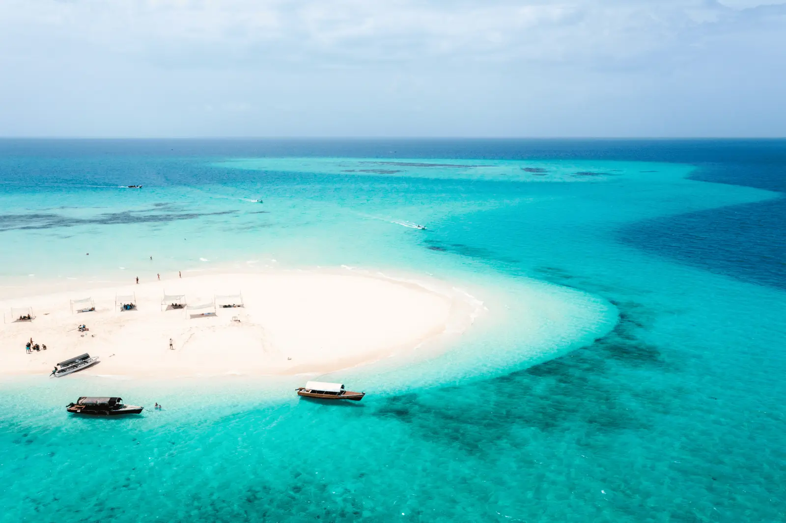 Curving tip of Nakupenda Sandbank with shade shelters and two wooden boats anchored in turquoise water