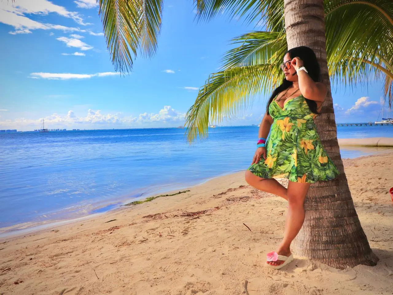 Woman in a green floral dress leaning against a palm tree on a sandy beach on Isla Mujeres