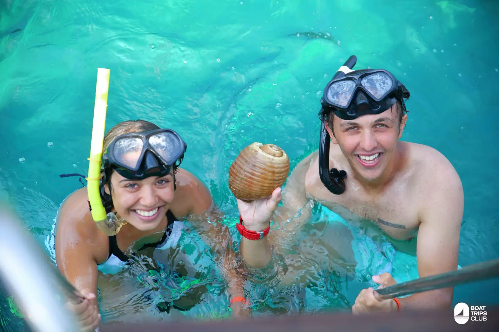 A smiling couple in snorkel masks shows off a seashell in the water beside the catamaran