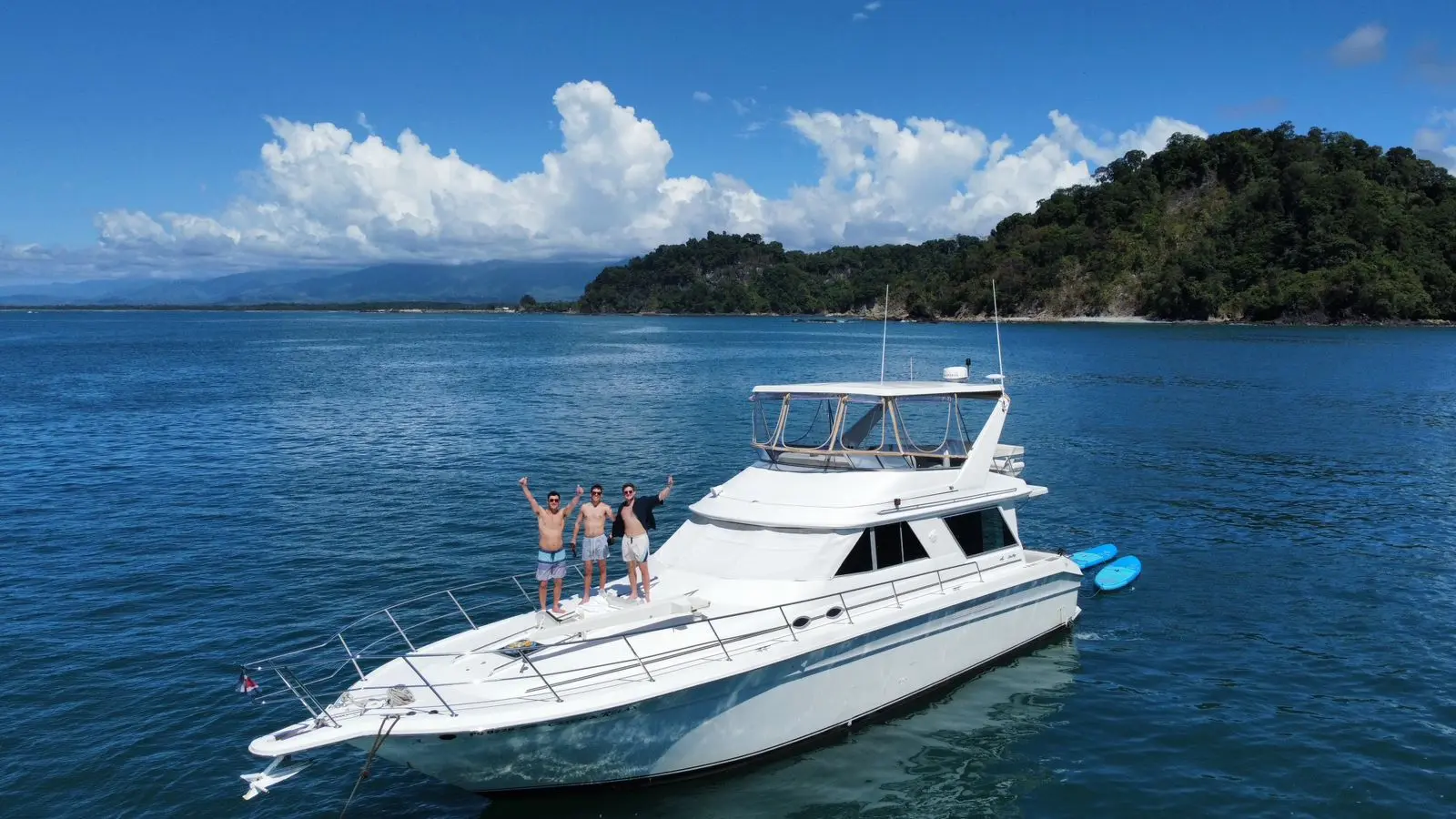 Three guests waving from the bow of a flybridge motor yacht anchored off the forested Manuel Antonio coast