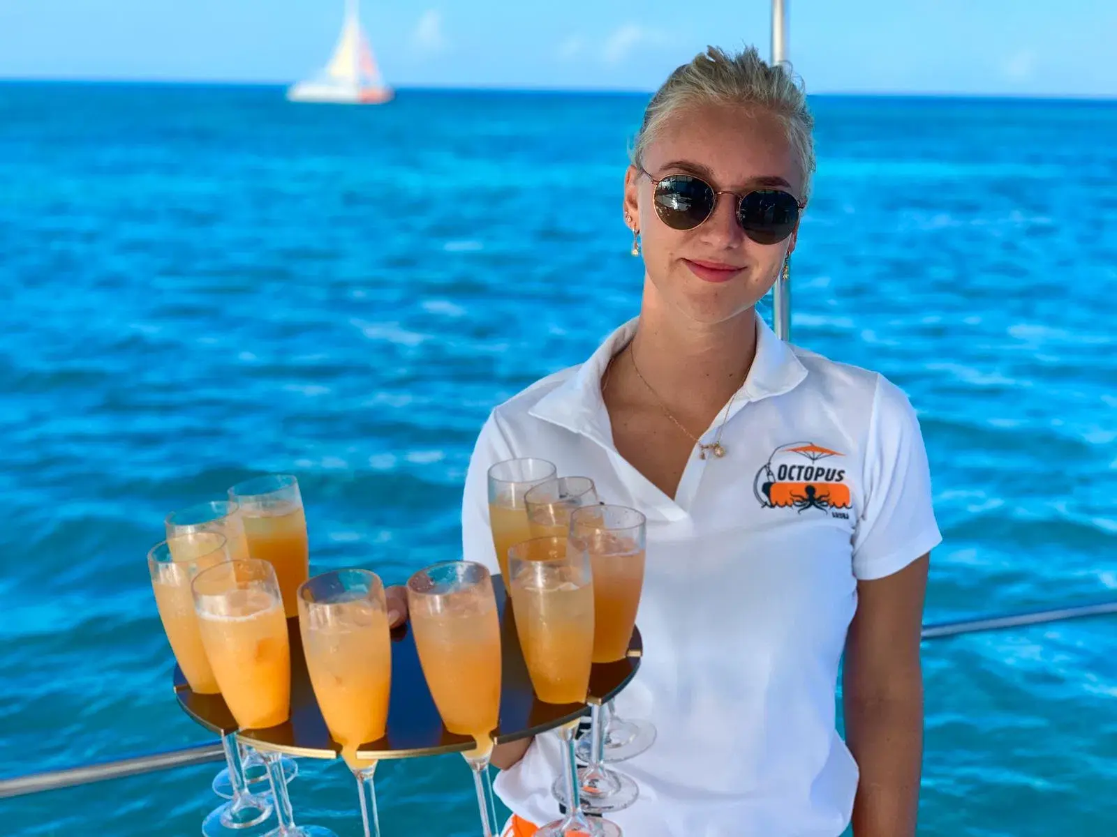 Crew member holding a tray of mimosas on the catamaran deck, a sailboat on the horizon