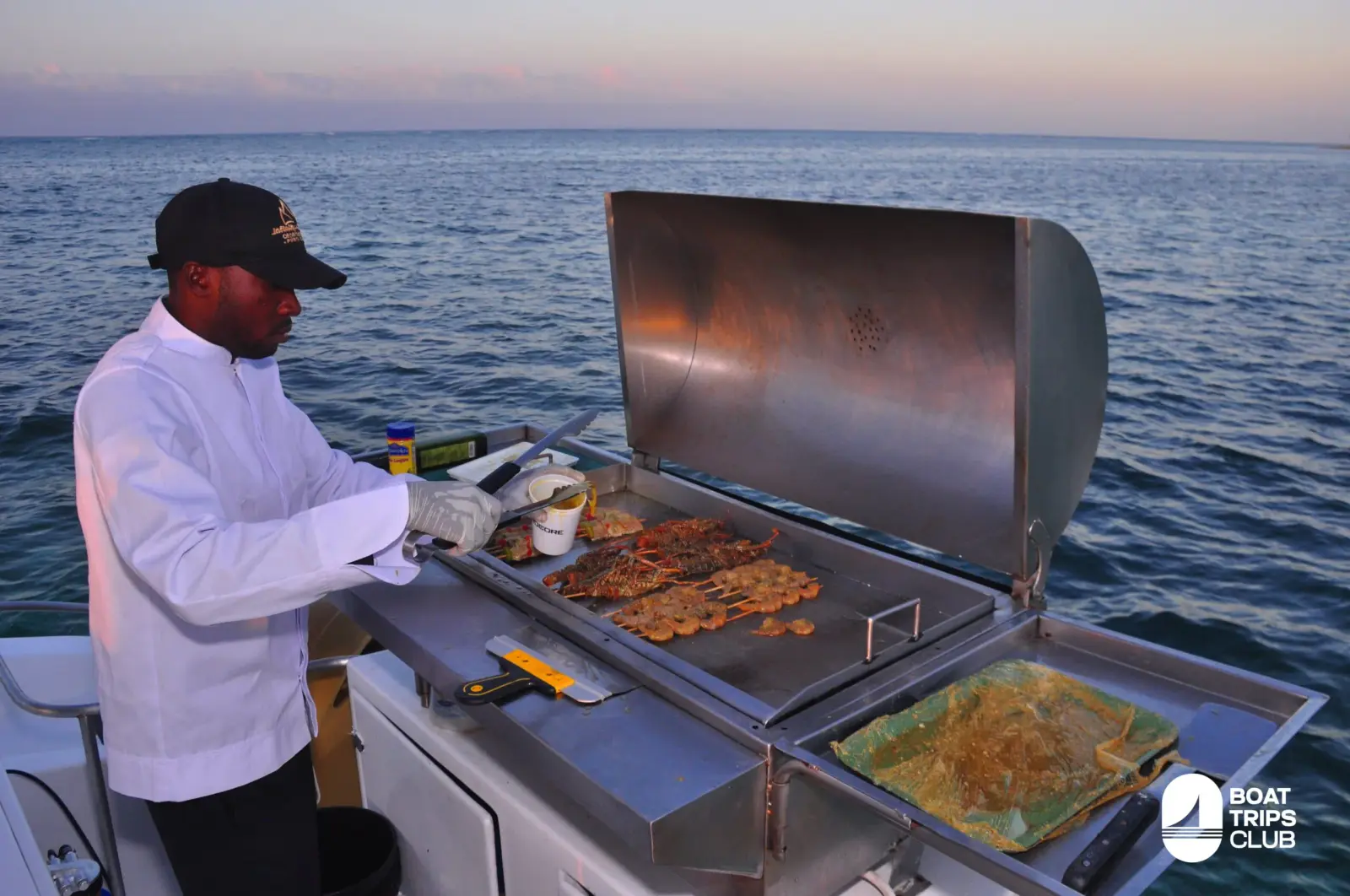 A crew member in a chef's jacket grills meat on a stainless steel barbecue on deck at dusk