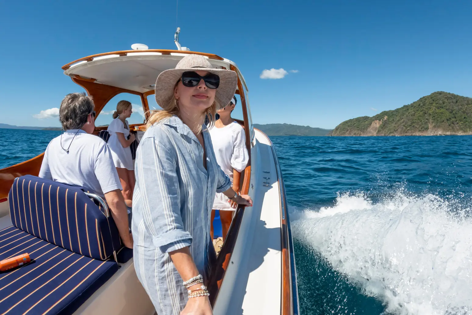 Woman in a sun hat standing in the cockpit of a picnic boat as it runs past a green headland, guests behind
