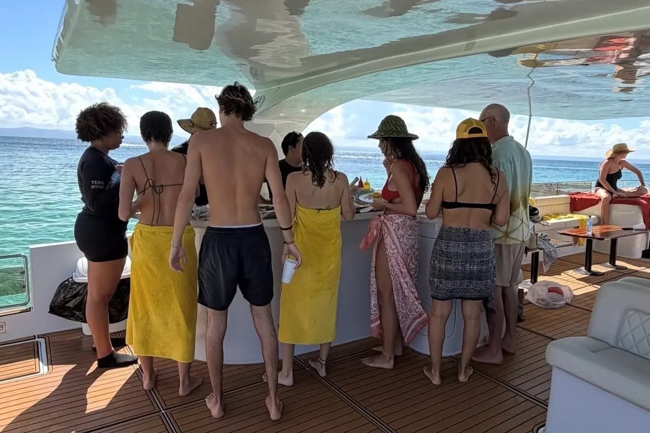 Guests in swimsuits and beach towels lining up at the counter on a power catamaran's shaded deck