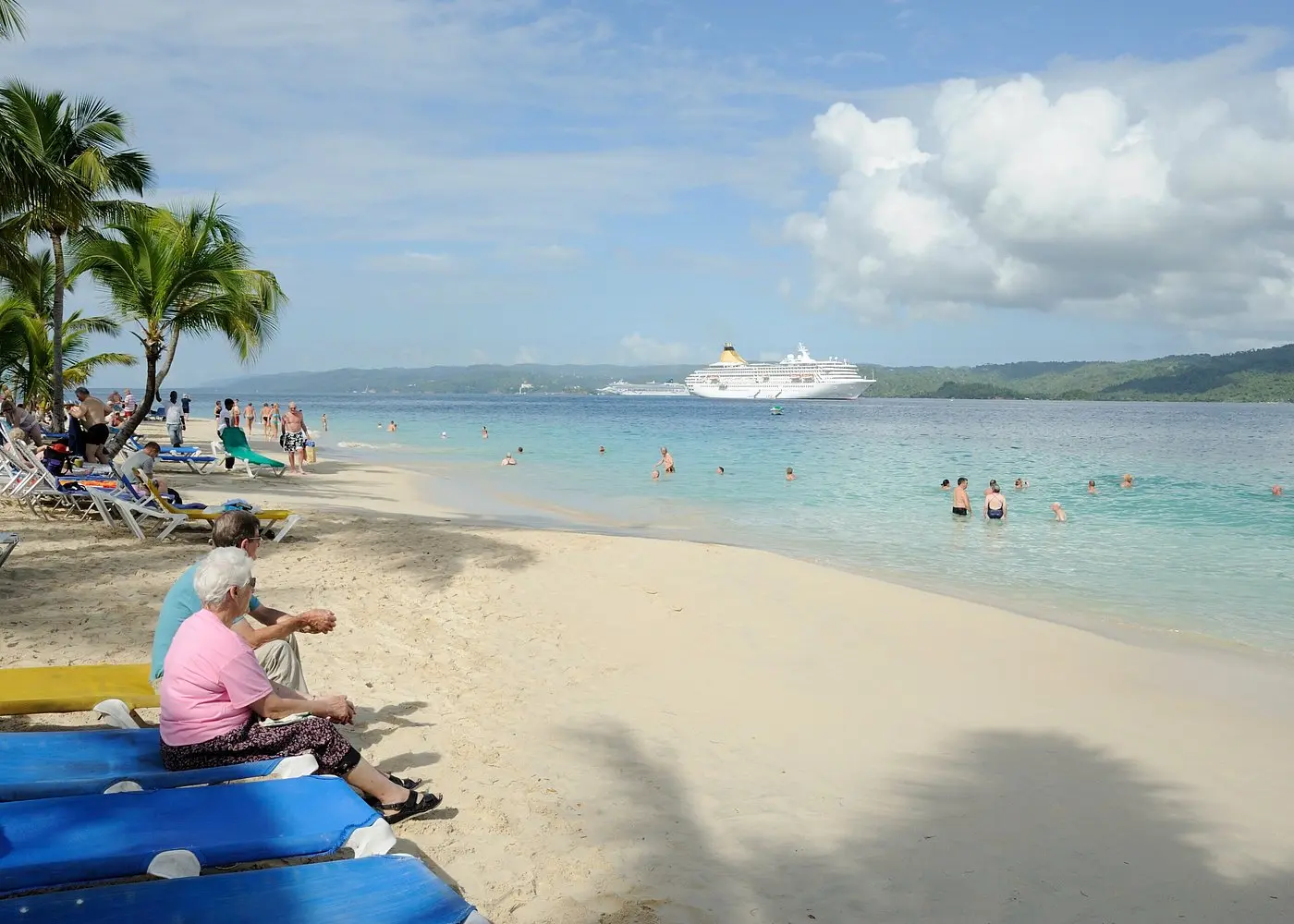 Beachgoers on blue loungers by the turquoise water of Cayo Levantado, a cruise ship anchored in the bay