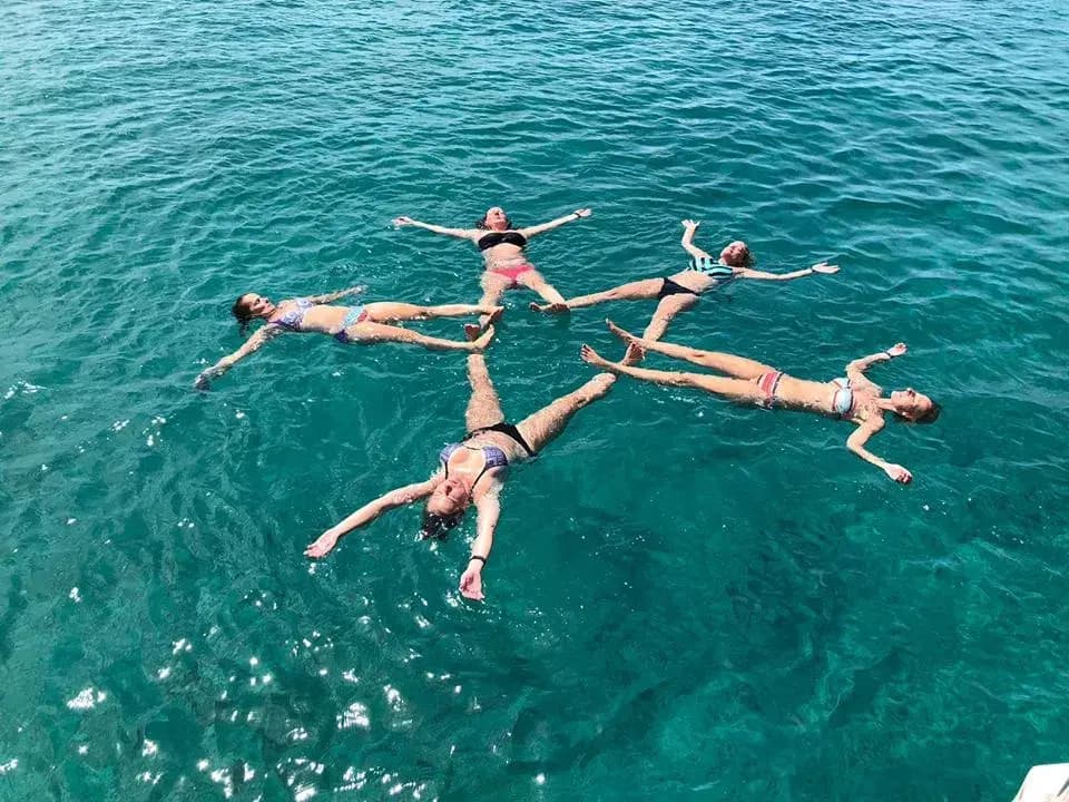 Five women floating on their backs in a star shape in clear turquoise water off Aruba