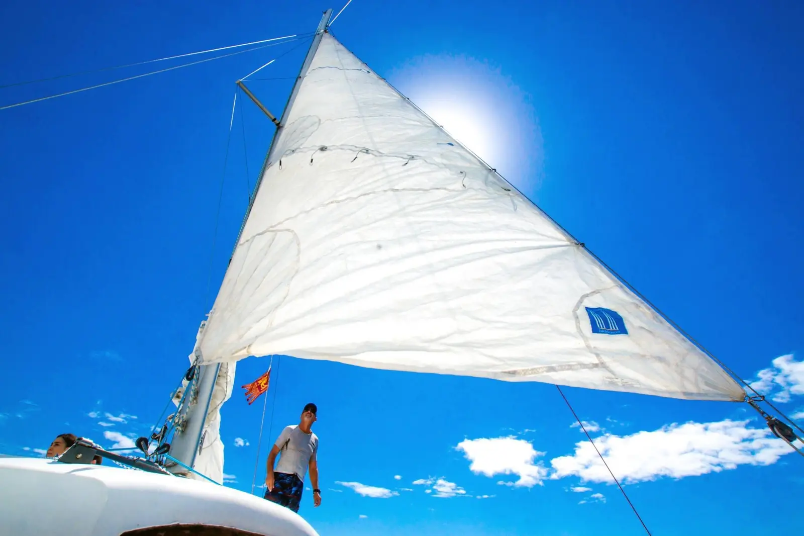Billowing white sail against a sunlit blue sky as a man stands on the catamaran's deck