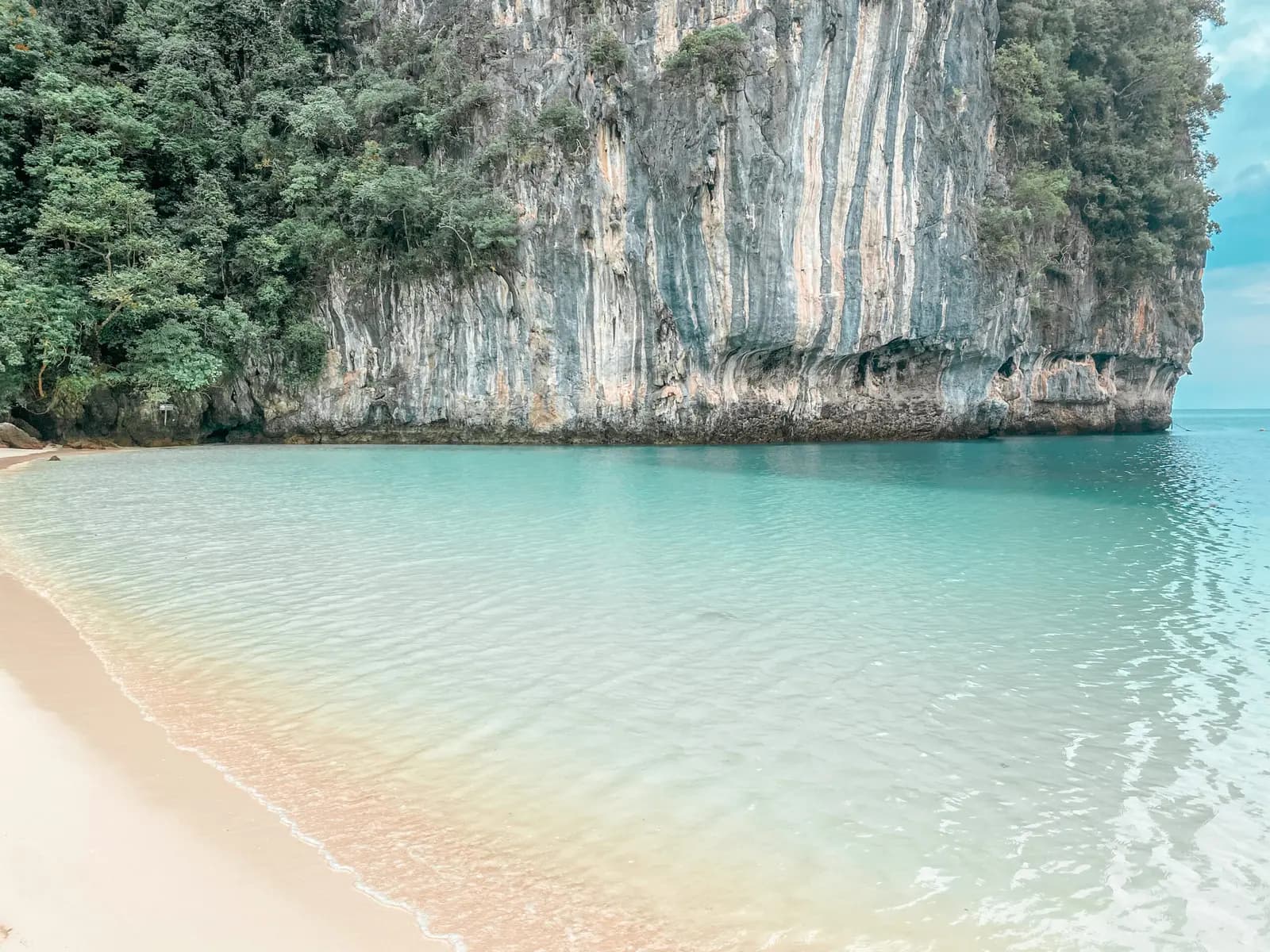 Empty curve of pale sand and glassy shallows beneath a sheer limestone cliff on Hong Island