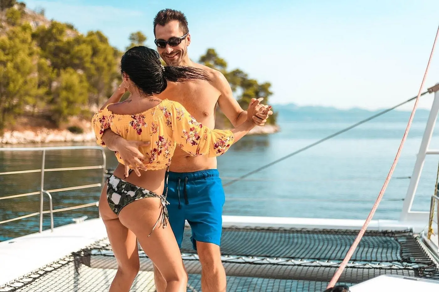 Couple dancing on the catamaran's foredeck by the trampoline net, a wooded Samaná coast behind