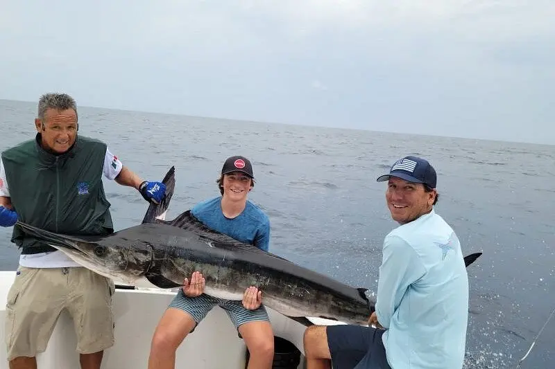 A man in a green vest and two anglers in caps holding a large marlin on a sportfishing boat