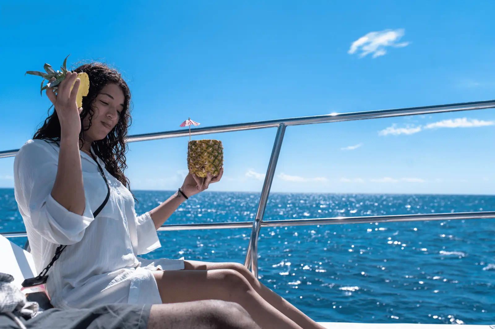 Woman in a white cover-up holding a pineapple cocktail by the rail above sparkling blue sea