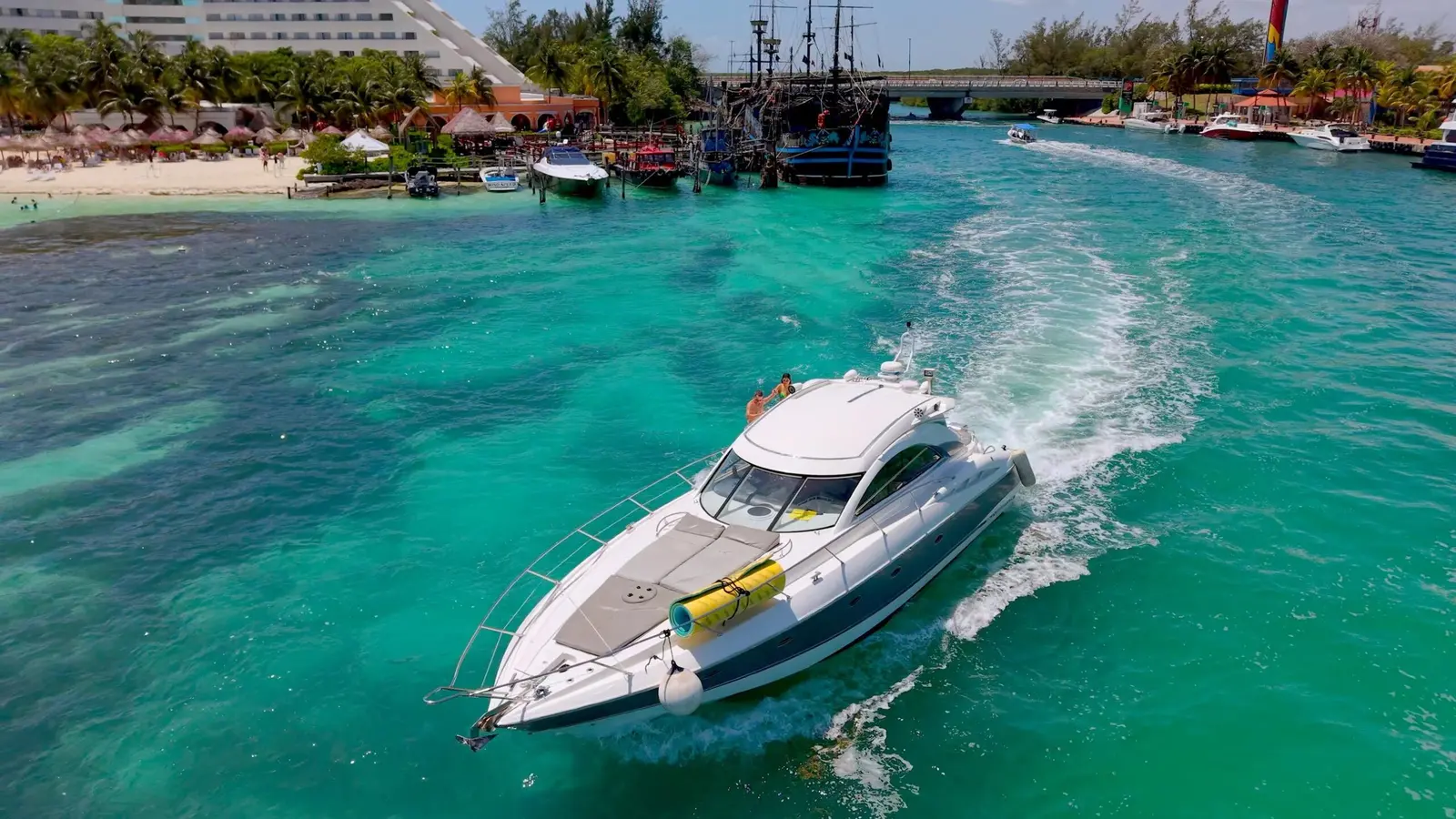 Motor yacht cruising over turquoise water past a beach and a moored pirate-style ship in Cancún