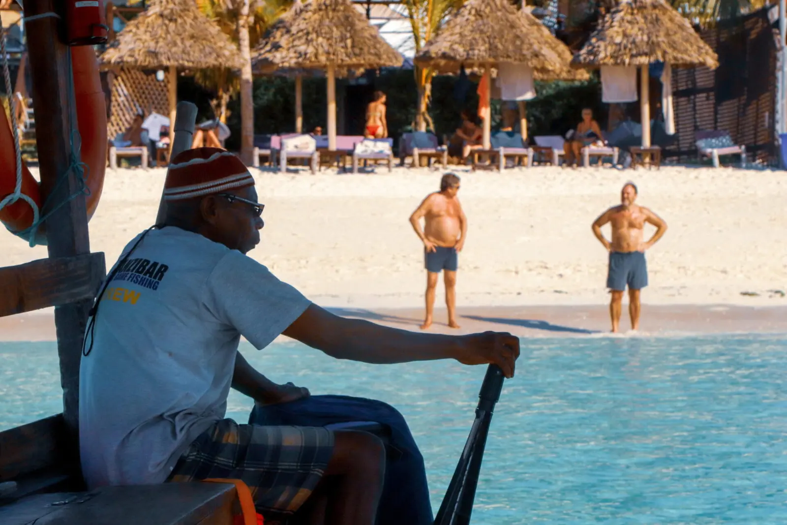 Crew member in a knit cap steering the dhow toward a Zanzibar beach lined with thatched umbrellas
