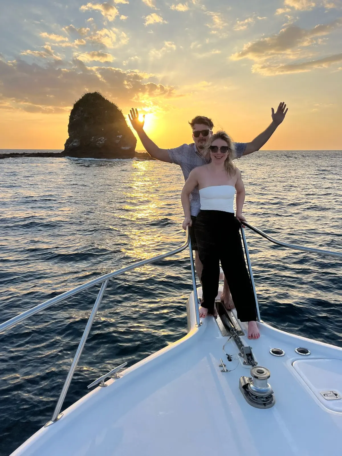 Couple posing on the yacht's bow at sunset beside a towering sea rock off Costa Rica