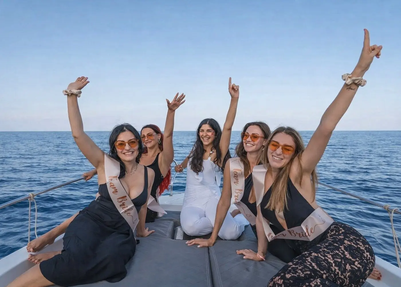 Five women in bachelorette sashes cheering with raised arms on the catamaran's padded bow in Bacalar