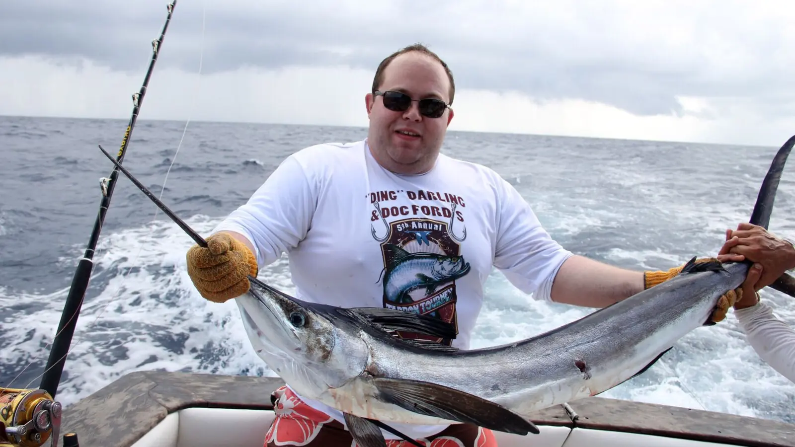 Angler in a white T-shirt holding a billfish beside the churning wake of a sportfishing boat