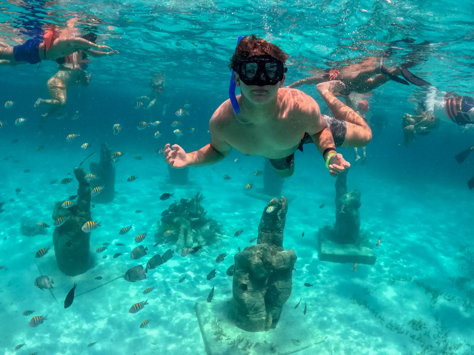 Snorkeler in a mask swimming among fish above underwater sculptures on the sandy seabed