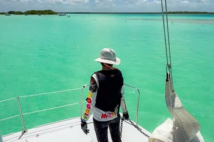 Crew member in a sun hat and gloves at the bow of a catamaran overlooking shallow Bacalar Lagoon