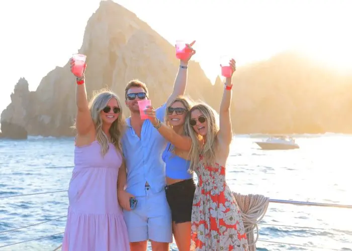 Four friends raising pink cocktails on a catamaran deck with sunlit Land's End rocks behind