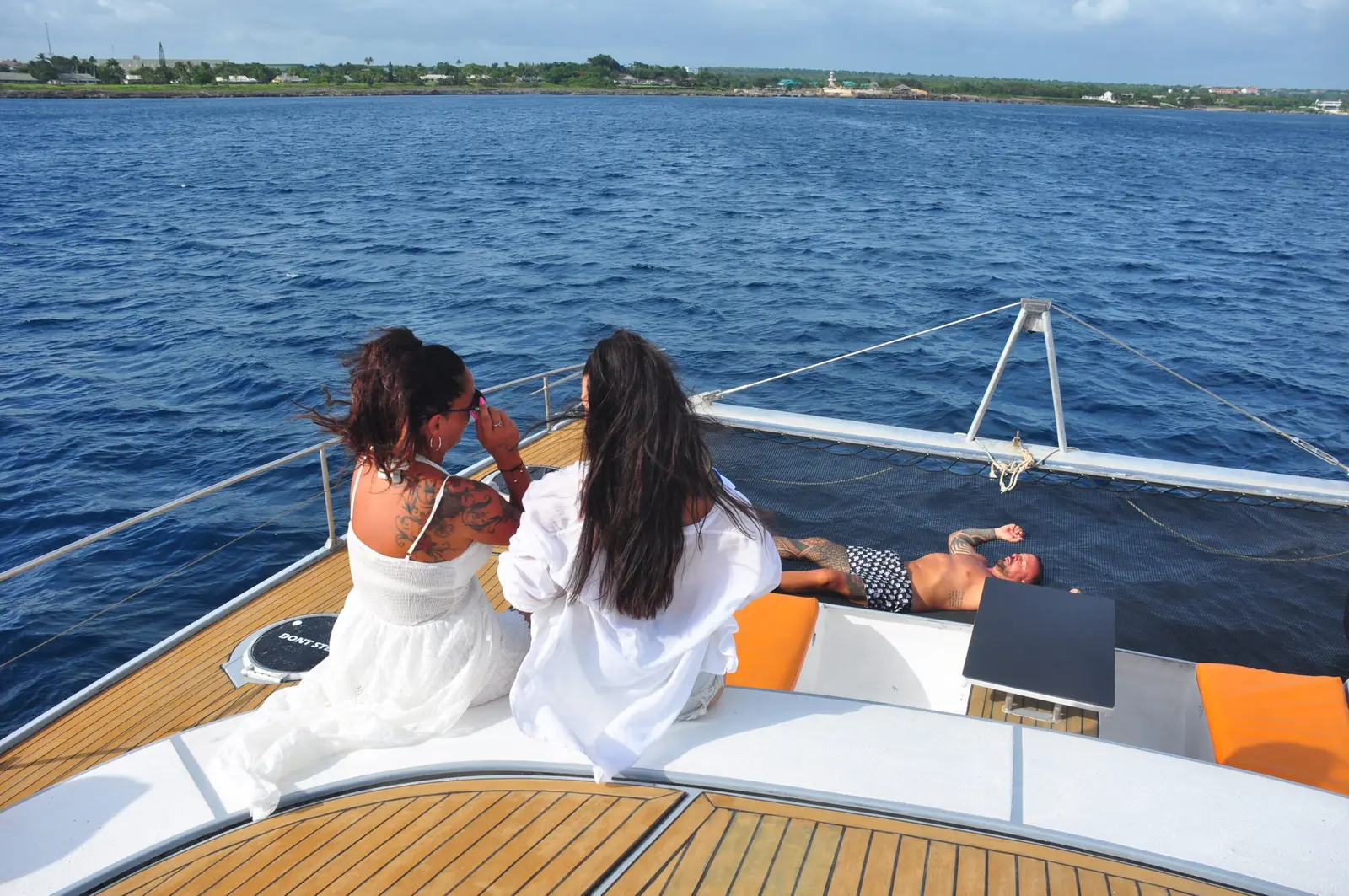 Two women sit at the catamaran's bow gazing across deep blue water toward a distant shore