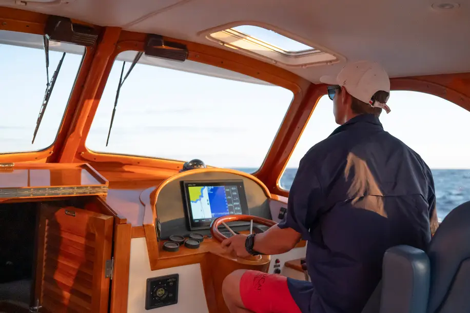 Captain steering the yacht from a varnished wood helm with a chartplotter screen and open sea ahead