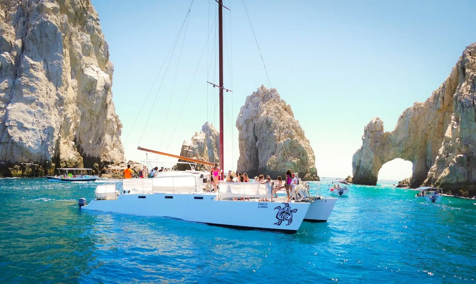 Sailing catamaran with guests aboard anchored in turquoise water beside El Arco