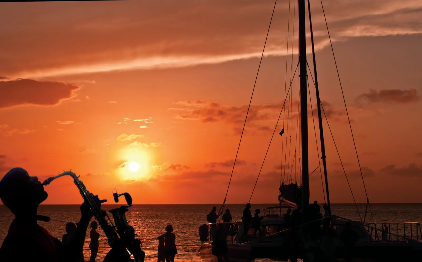 Saxophonist playing as guests stand silhouetted on a sailing catamaran's deck against an orange sunset
