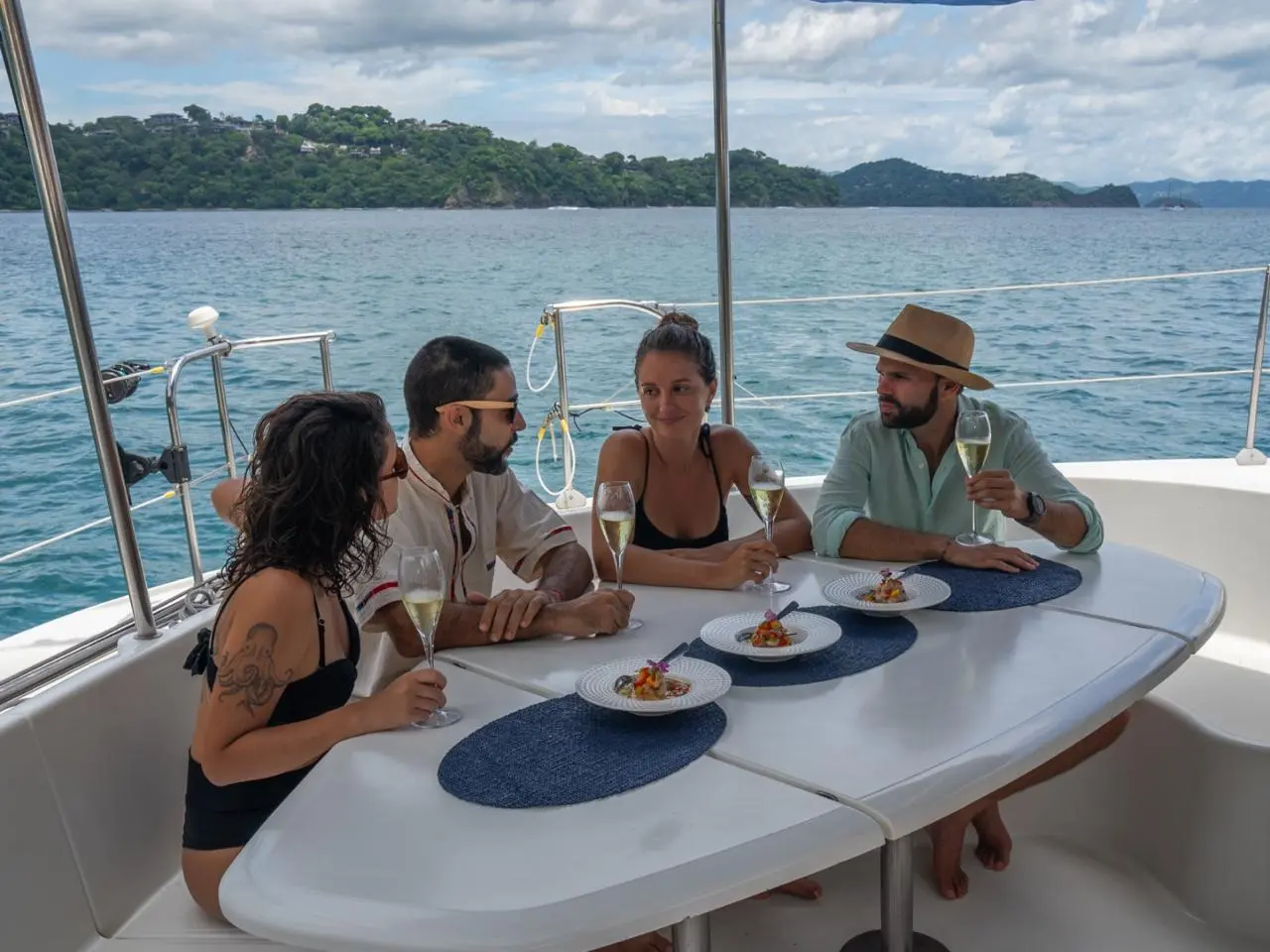 Four guests share a plated lunch with sparkling wine at the deck table of the catamaran