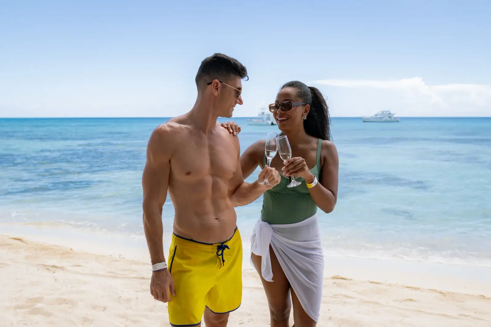 A couple clinks wine glasses on a white sand beach with boats anchored offshore