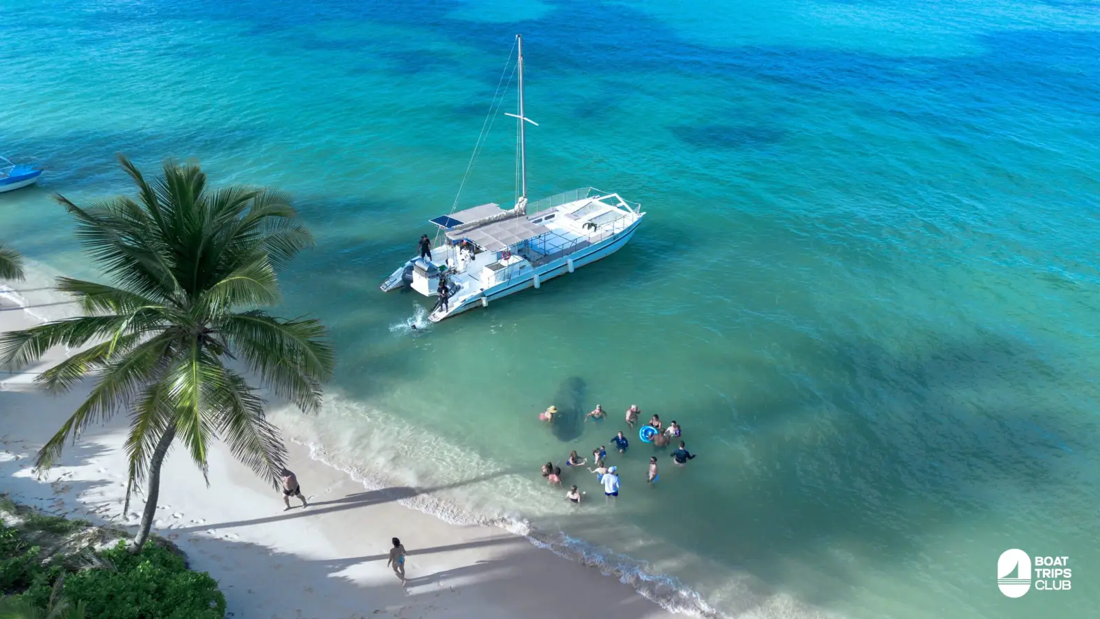 Guests wade in the shallows between a sailing catamaran and a palm-shaded white sand beach in Bavaro